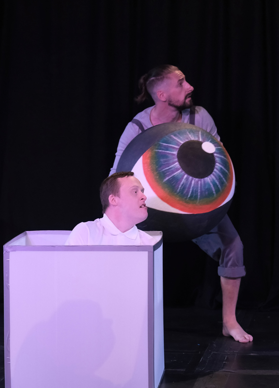 A photograph of two male dancers performing SENse. One is sat in a white box on the floor and the other is holding a giant eye.