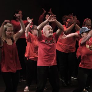 A photograph of participants from our Everybody Dance Chorley session. They are on stage, all wearing red, with their arms raised while dancing. 