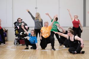 An inclusive team of dancers of all different abilities are posing with their arms lifted