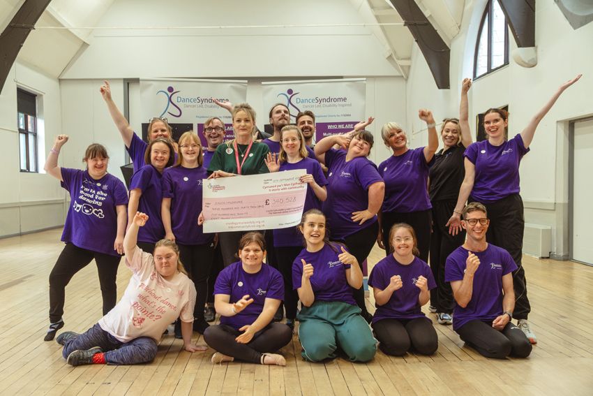 A photograph of Beth Cox from The National Lottery Community Fund with the DanceSyndrome DS Collective performance team. They are holding a giant cheque and cheering.
