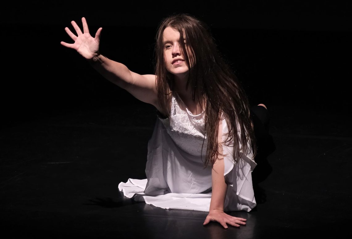 A dancer in a white dress is kneeling on the floor reaching her arm out, against a black background.