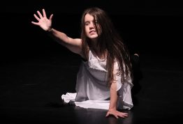 A dancer in a white dress is kneeling on the floor reaching her arm out, against a black background.
