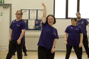 A female dancer is stood at the front with her arms raised. She has other dancers in spaces behind her. 