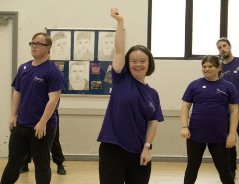 A female dancer is stood at the front with her arms raised. She has other dancers in spaces behind her.
