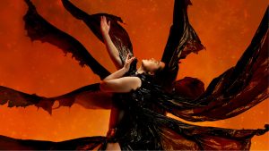 A photograph of a dancer wearing a black costume which is blowing in the wind, set against an orange background.