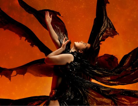 A photograph of a dancer wearing a black costume which is blowing in the wind, set against an orange background.