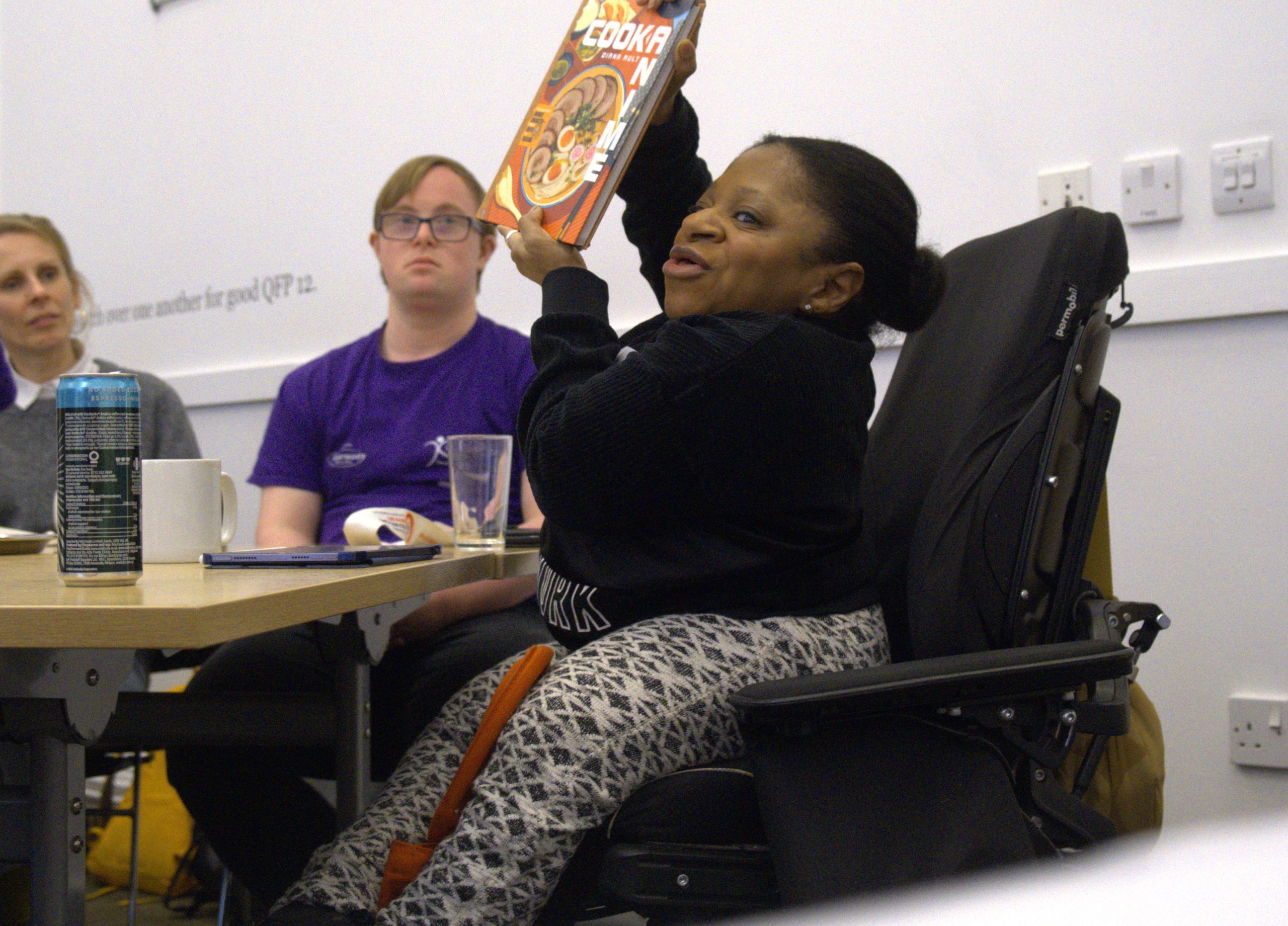 An Artist who is a wheelchair user is holding up a book to show to the other delegates, who are looking on.