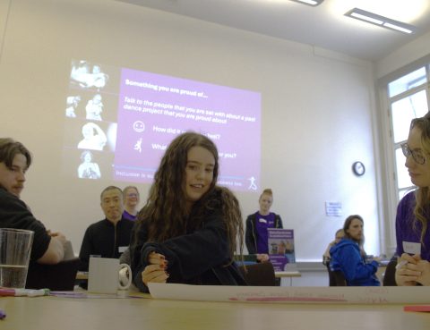 A photograph of an Artist with a learning disability at the Artists with Disabilities Sharing and Learning event. There are people sat at the table with her and a projector screen in the background