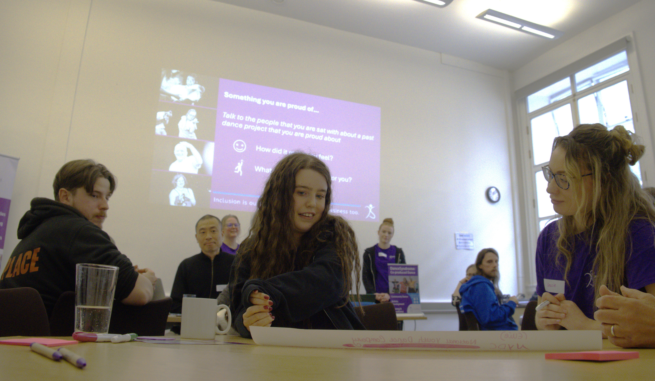A photograph of an Artist with a learning disability at the Artists with Disabilities Sharing and Learning event. There are people sat at the table with her and a projector screen in the background