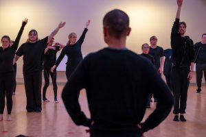 A choreographer watching dancers move around a studio with their arms raised